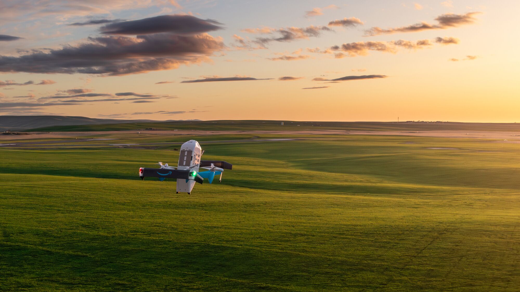 Official Amazon photograph of a Prime Air delivery drone hovering low over open green grassland at sunset, its rotors blurred, with a long flat horizon behind it.