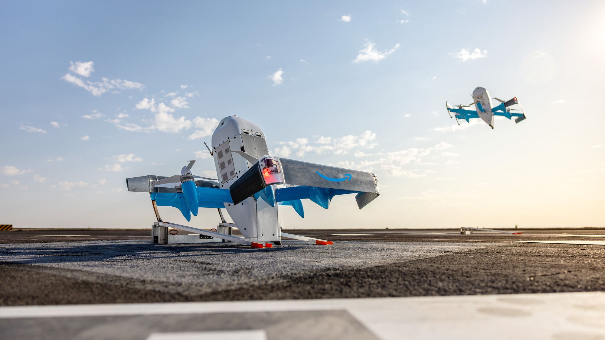 Official Amazon photograph of two MK30 Prime Air delivery drones, hexagonal white and grey airframes with six enclosed rotor housings, on a pale surface in bright studio lighting.