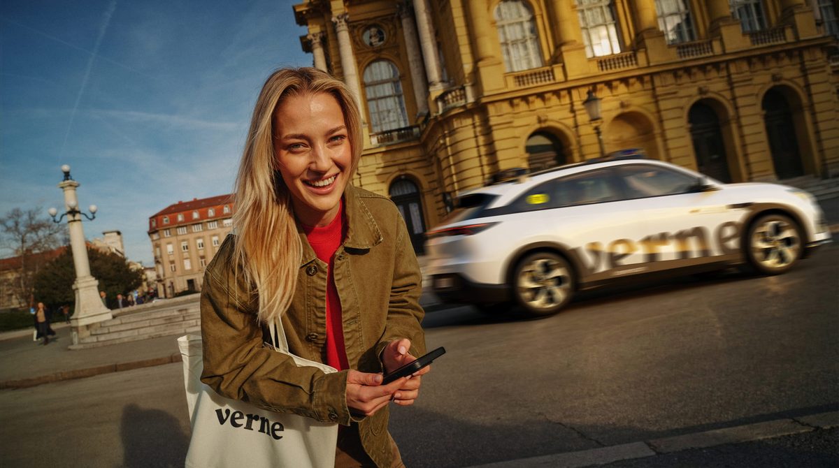 A Verne-branded white robotaxi drives past the Croatian National Theatre in Zagreb while a passenger holding a phone stands in the foreground, from Verne's press material for Europe's first commercial robotaxi service.