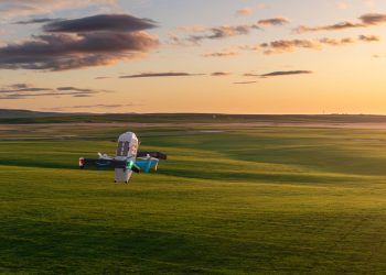 Official Amazon photograph of a Prime Air delivery drone hovering low over open green grassland at sunset, its rotors blurred, with a long flat horizon behind it.