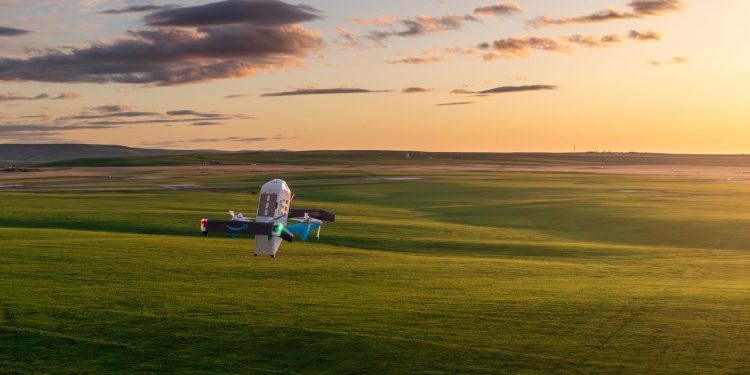Official Amazon photograph of a Prime Air delivery drone hovering low over open green grassland at sunset, its rotors blurred, with a long flat horizon behind it.