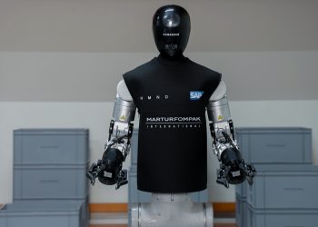The HMND 01 Alpha humanoid robot standing in an industrial facility in a black vest branded HMND, SAP and Martur Fompak, with two arms ending in parallel grippers and grey logistics totes stacked around it.
