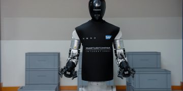 The HMND 01 Alpha humanoid robot standing in an industrial facility in a black vest branded HMND, SAP and Martur Fompak, with two arms ending in parallel grippers and grey logistics totes stacked around it.