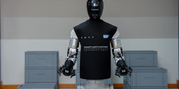 The HMND 01 Alpha humanoid robot standing in an industrial facility in a black vest branded HMND, SAP and Martur Fompak, with two arms ending in parallel grippers and grey logistics totes stacked around it.