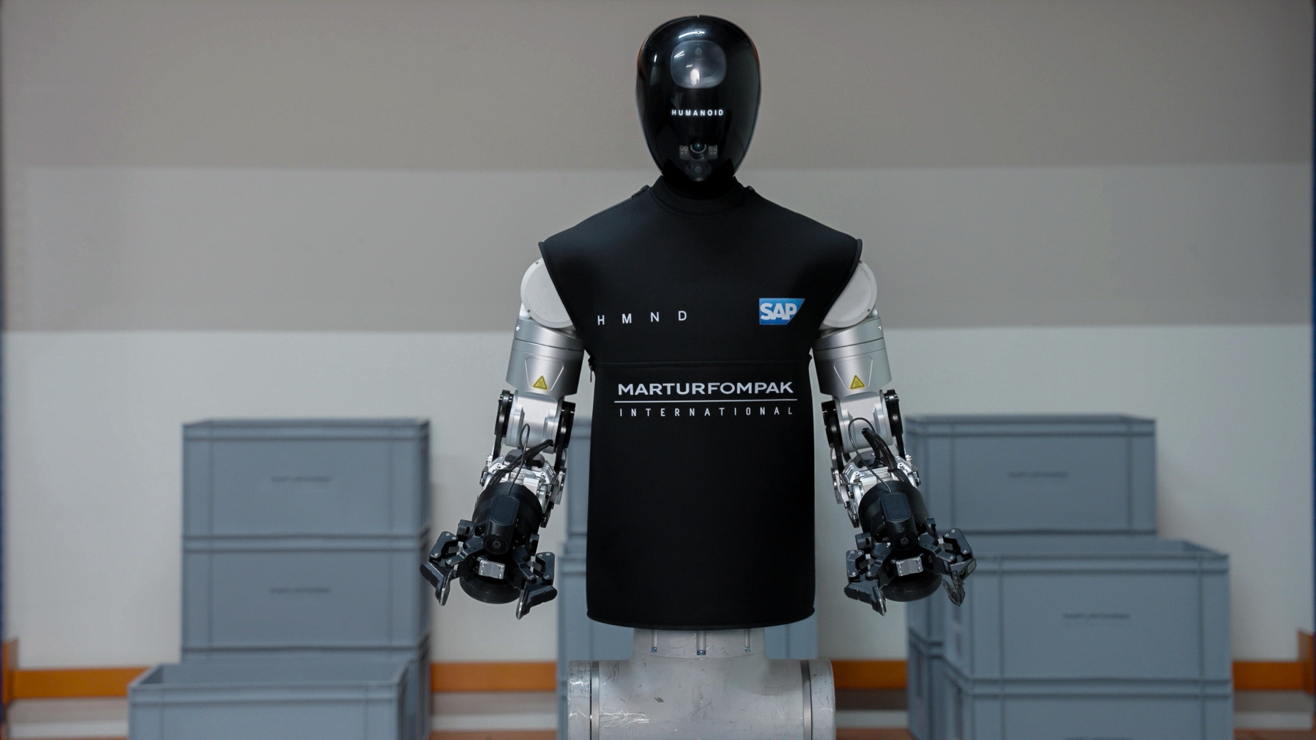 The HMND 01 Alpha humanoid robot in a black vest branded HMND, SAP and Martur Fompak, standing between grey logistics totes with both arms ending in black parallel grippers.