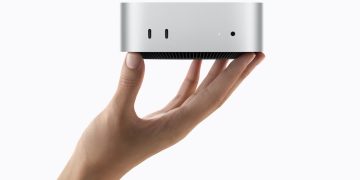 Official Apple product photograph of the 2026 Mac mini, a small square silver aluminium desktop computer with rounded corners and a dark circular base, shown at a three quarter angle against a plain light background.