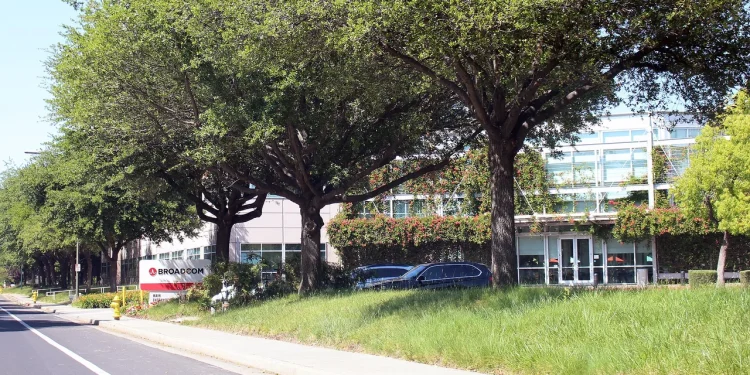 Broadcom Inc. headquarters in San Jose, California: the Broadcom sign by the roadside and an office building with a glass entrance behind a row of trees