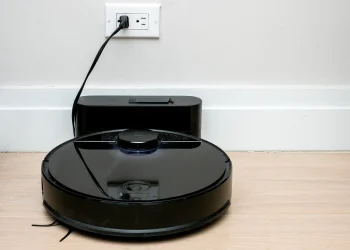 Black Roborock robot vacuum cleaner docked at its charging base against a white wall on a wooden floor