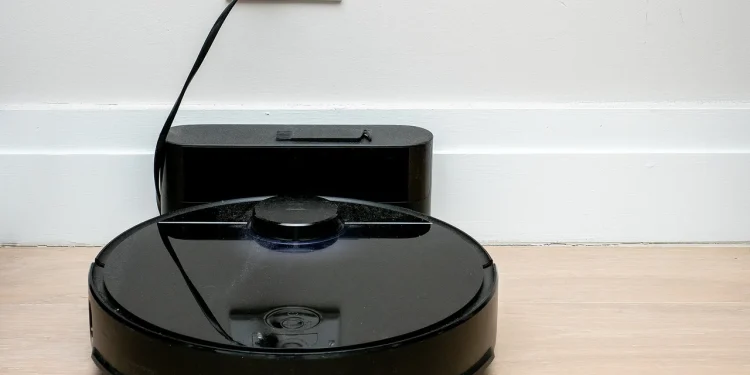 Black Roborock robot vacuum cleaner docked at its charging base against a white wall on a wooden floor