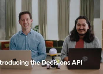 OpenAI's announcement video still for the Decisions API: two presenters at a table with laptops and the caption 'Introducing the Decisions API'