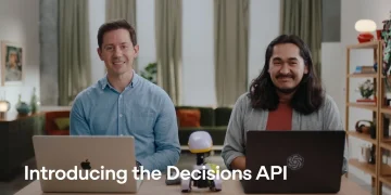 OpenAI's announcement video still for the Decisions API: two presenters at a table with laptops and the caption 'Introducing the Decisions API'