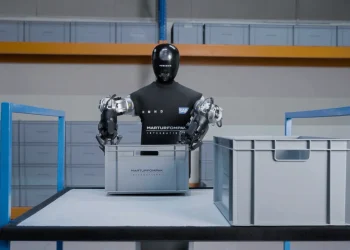 The HMND 01 Alpha humanoid robot with a black head and two gripper arms handling a gray crate in a warehouse during a pilot with SAP and Martur Fompak