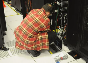 Technician in a red plaid shirt crouching beside an open server rack with a laptop on the floor and network cables hanging from the rack