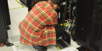 Technician in a red plaid shirt crouching beside an open server rack with a laptop on the floor and network cables hanging from the rack