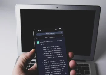 A hand holding a smartphone that shows a ChatGPT conversation, in front of a laptop with a dark screen