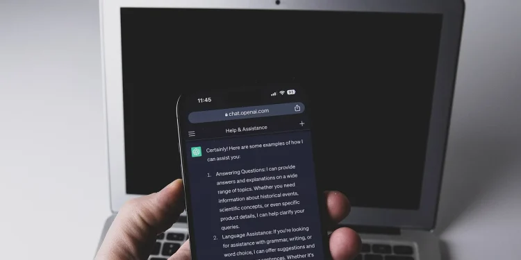 A hand holding a smartphone that shows a ChatGPT conversation, in front of a laptop with a dark screen
