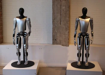 Two Tesla Optimus humanoid robots with silver torsos and black heads standing on display plinths in front of a white brick wall