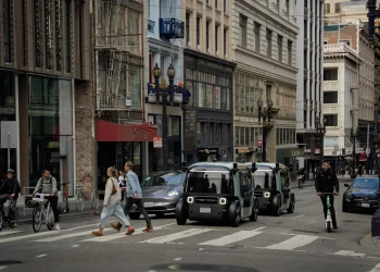 Two Zoox robotaxis without steering wheels driving through a busy downtown street crossing past pedestrians, cyclists and a scooter rider