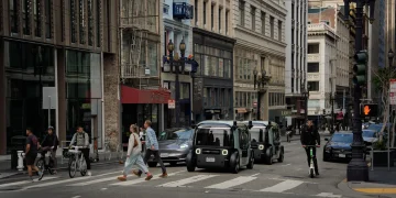 Two Zoox robotaxis without steering wheels driving through a busy downtown street crossing past pedestrians, cyclists and a scooter rider